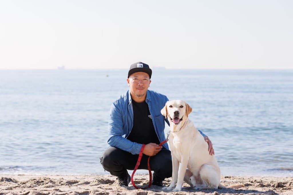 理事長の近藤が砂浜で黄色いラブラドールレトリバーのそばにしゃがみ、リードを握っている様子。背景は海と青空。